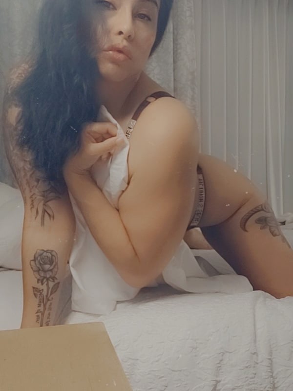 A woman with long dark hair sitting on a bed, wearing lingerie and a towel, with tattoos visible on her arms.