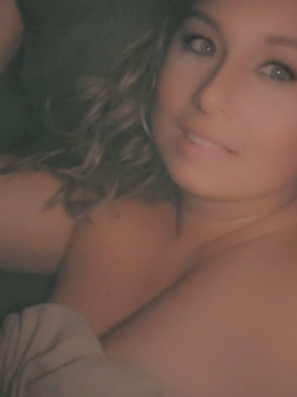 Close-up portrait of a smiling woman with wavy hair lying on a bed, wearing a light top.