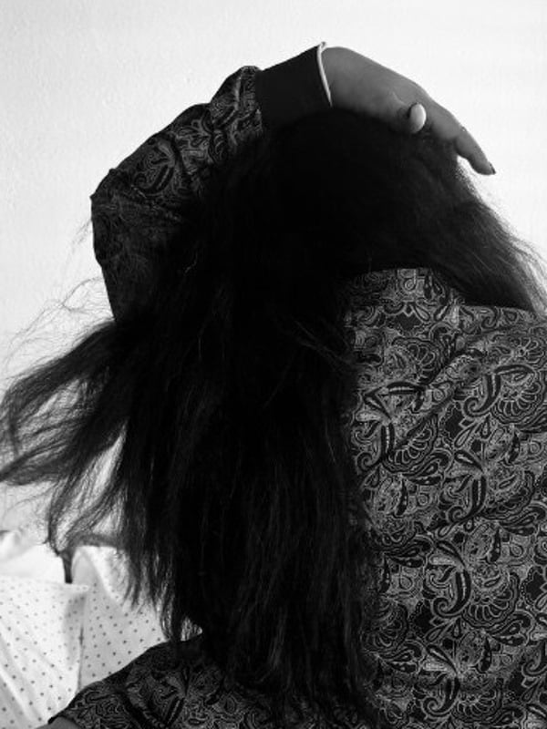 Black and white photo of a woman from behind, with long dark hair cascading over her shoulders, standing near a window with blinds.