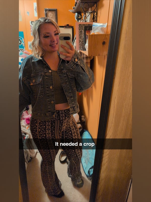 A woman with blonde hair takes a selfie in a mirror, wearing a denim jacket over a green top and patterned pants, smiling at the camera in a cozy room.