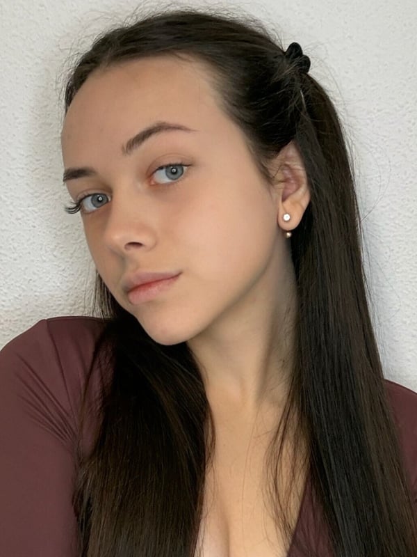 Close-up portrait of a young woman with long dark hair in pigtails, subtle makeup, wearing small earrings and a maroon top, gazing sideways against a plain background.