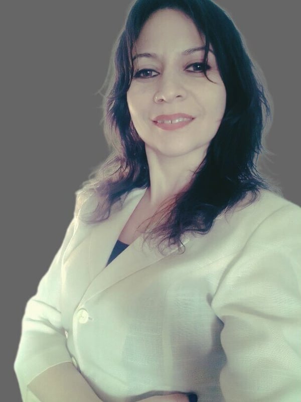 A woman with wavy dark hair smiling warmly while wearing a white blazer against a gray background.