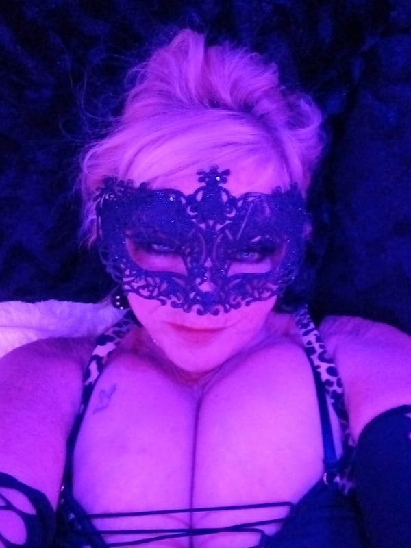 Woman with blonde hair wearing a lace mask and black top, reclining on a dark surface with purple lighting.
