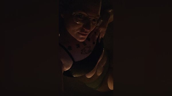A woman with dark hair posing closely in dim lighting, wearing a light-colored top with tattoos visible on her shoulder.