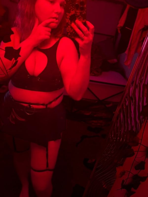 A woman taking a selfie in a low-cut black top and skirt with garter straps, standing in a red-lit room.