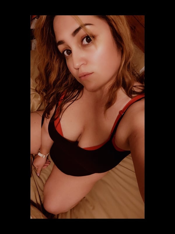 Close-up selfie of a woman with wavy brown hair, wearing a red bra top, posing indoors.
