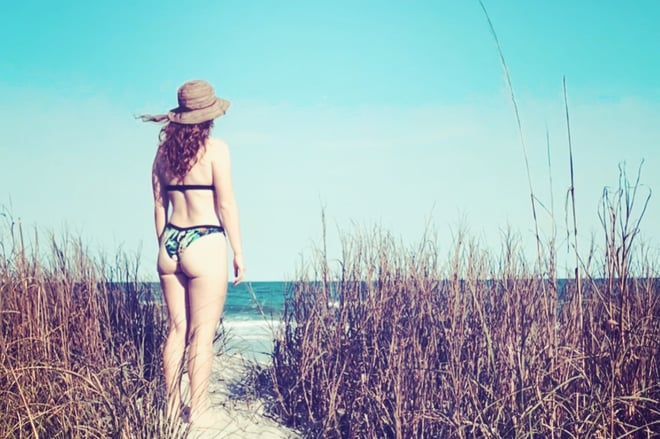 A woman in a bikini and hat stands on a beach path surrounded by tall grasses with the ocean in the background.