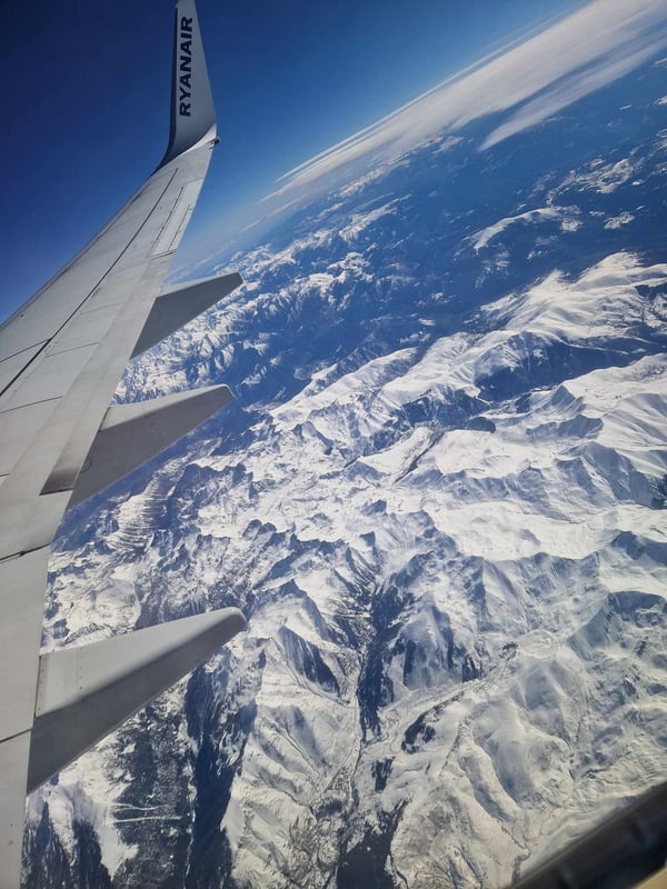 Aerial view from an airplane window showing a Ryanair plane wing over snow-capped mountains and clouds.