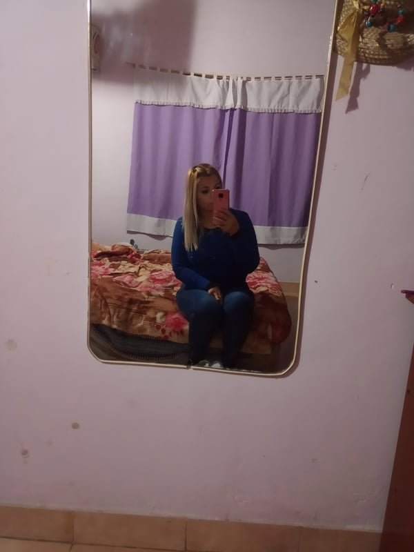 Young woman with long blonde hair sitting on a bed in front of a mirror, wearing a blue sweater and pants in a cozy bedroom with purple curtains.