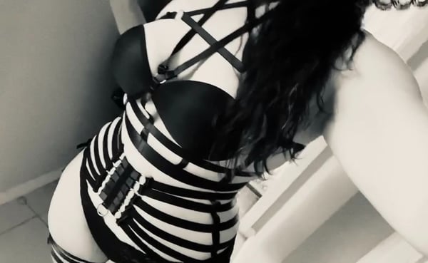 Woman posing in black harness attire with long dark hair in an indoor setting.