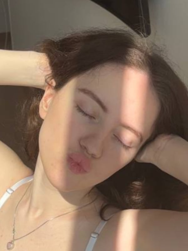 Close-up of a young woman with long brunette hair making a playful kiss face while wearing a white bra, with natural light illuminating her face.