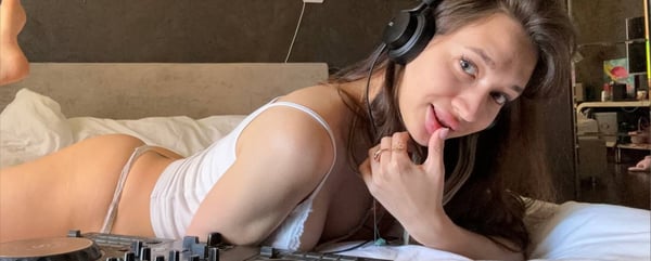 A woman lying on a bed wearing headphones and a white top, with DJ equipment nearby in a cozy indoor setting.