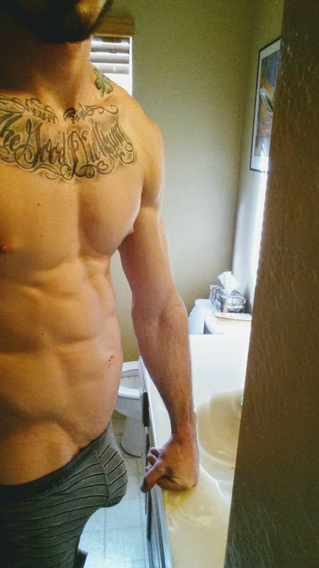 A muscular man with tattoos on his chest and neck stands in a bathroom, wearing gray underwear and posing confidently near a sink.