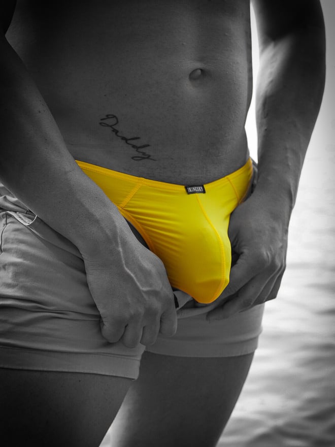 Close-up of a shirtless man in yellow underwear and gray shorts, standing confidently with tattoos visible on his abdomen.