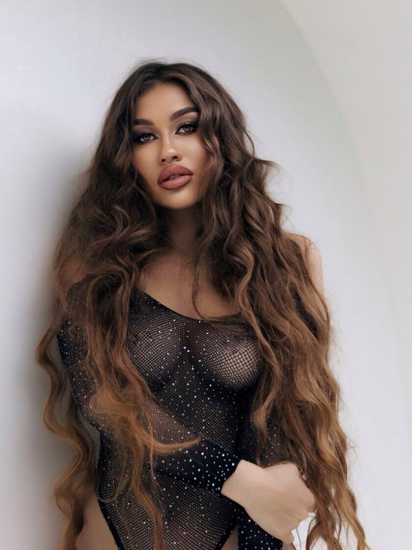 A woman with long wavy brown hair stands against a white curved wall, dressed in a sheer sparkling black bodysuit.
