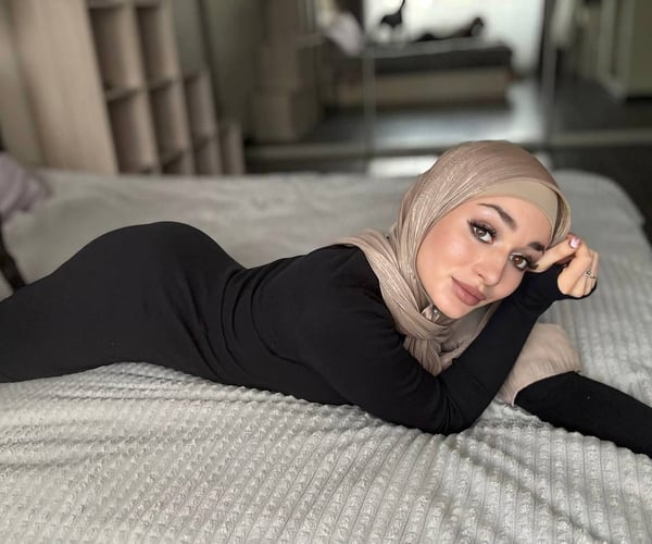 A woman wearing a beige hijab and black form-fitting outfit reclines on a bed in a modern bedroom with shelves and mirrors in the background.
