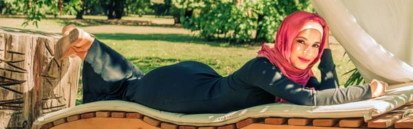A woman in a pink hijab and black outfit reclining on a wooden lounge chair in a lush green outdoor setting with draped fabric.