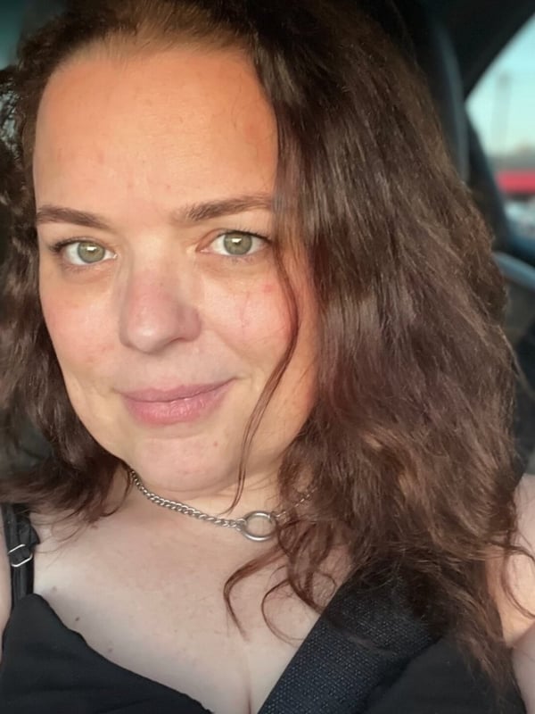 A woman with wavy brown hair and green eyes smiling in a car, wearing a black top and necklace.