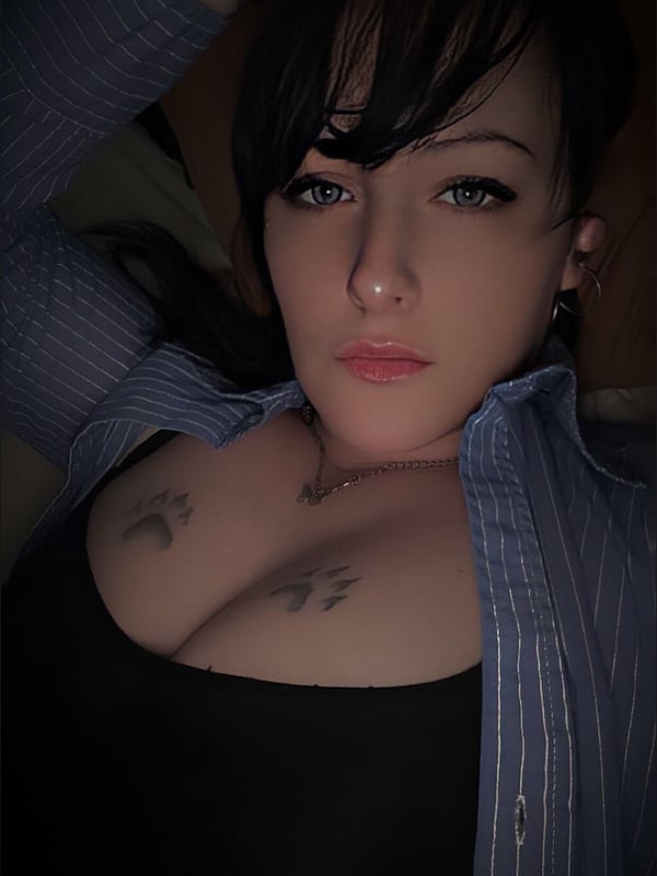 Close-up portrait of a woman with dark hair, tattoos on her chest, and piercings, wearing an open blue shirt over a black top.