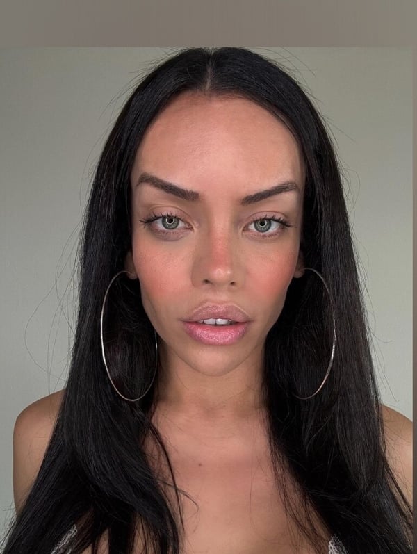 A person with long dark hair and hoop earrings posing against a plain background.