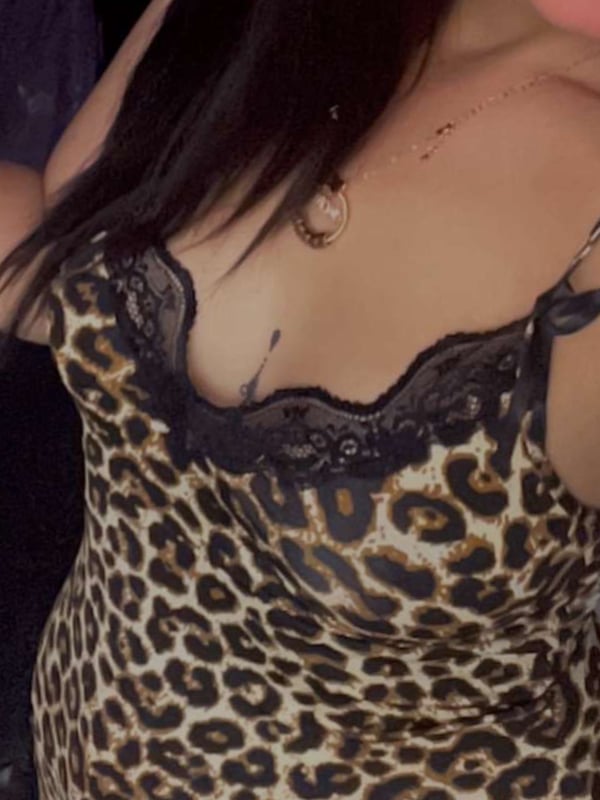 A woman with long dark hair wearing a lace-trimmed leopard print top, posing confidently indoors.