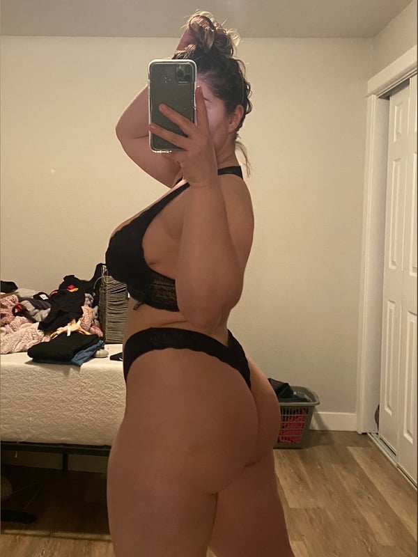 A woman taking a mirror selfie in a bedroom, wearing black lingerie and holding her phone.