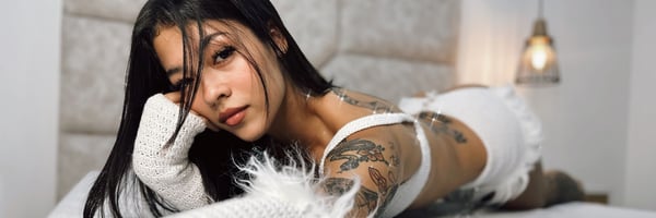 Young woman with long dark hair reclining on a bed in white lingerie, with tattoos visible on her arms and soft lighting from a bedside lamp.