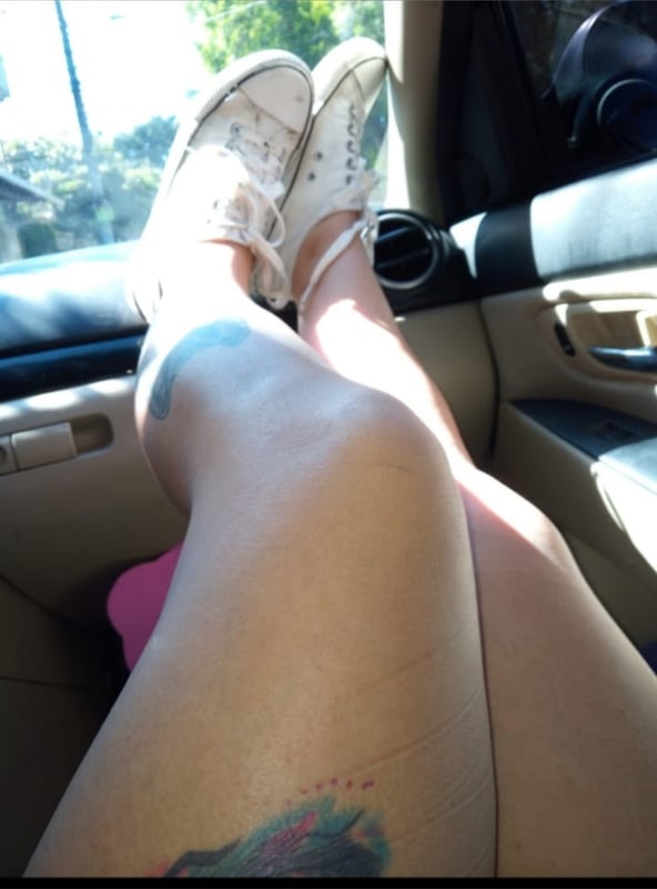 Person's legs in white sneakers propped up on a car dashboard with tattoos visible on the legs, inside a vehicle with natural light filtering through the window.