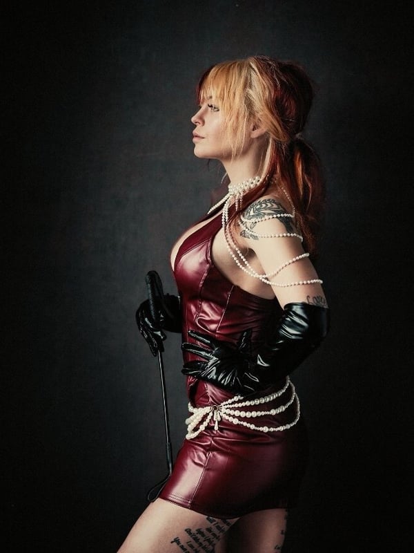 A woman in red latex outfit and black gloves holding a whip, posing confidently in a studio setting.