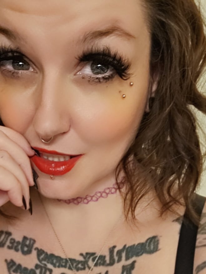 Close-up portrait of a woman with heavy makeup, piercings, tattoos, and a choker necklace, smiling playfully.