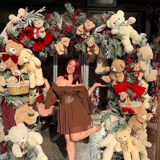 A young woman with red hair smiling and posing playfully in front of a large Christmas decoration featuring teddy bears, garlands, and wreaths.