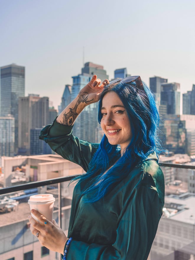 A smiling woman with blue hair and tattoos stands on a rooftop holding a coffee cup, with a city skyline in the background.