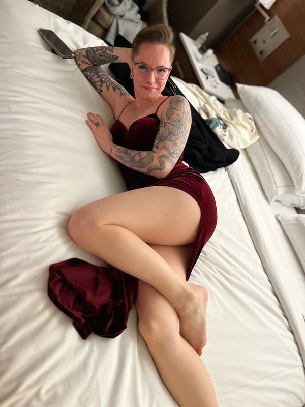 A woman with tattoos reclines on a bed in a maroon dress, smiling at the camera in a cozy hotel room.