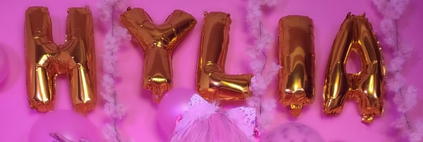 Decorative gold balloon letters spelling 'HYLIA' arranged with pink balloons and feathers on a pink background.