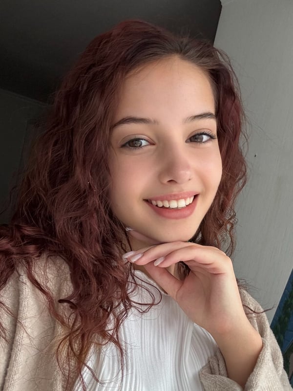 Young woman with wavy red hair smiling warmly, posing with hand on chin while wearing a white top and beige cardigan against an indoor background.