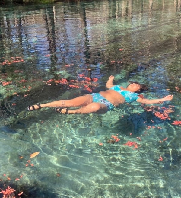 A woman relaxing in a clear natural spring surrounded by trees and colorful leaves, wearing a bikini and sandals.