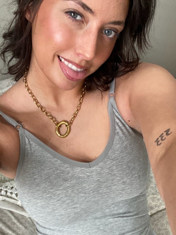 Young woman with wavy dark hair smiling at the camera in a gray tank top, gold chain necklace, and tattoos on her arm, posing indoors.