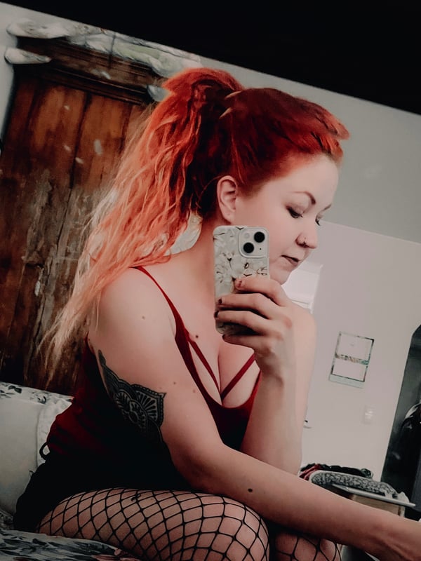 A woman with long red hair in a ponytail taking a mirror selfie in a tank top and shorts with fishnet stockings, holding a phone.