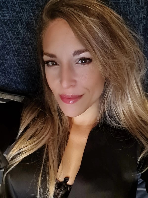 A woman with long blonde hair smiling confidently at the camera, dressed in a black top against a textured background.
