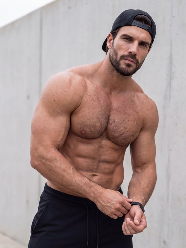 A muscular man with dark hair and a beard wearing a black cap, posing shirtless outdoors against a concrete wall.