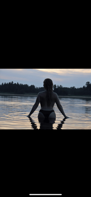 A woman standing by the water's edge at sunset, wearing a swimsuit bottom, with her back to the camera and arms extended towards the lake.