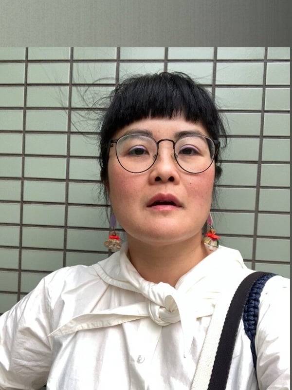 A woman with glasses and dark bangs wearing a white hooded top, standing against a green tiled wall.