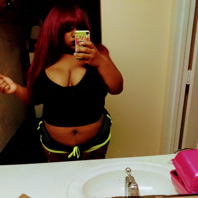 A woman with long red hair taking a selfie in a bathroom, wearing a black top and green-trimmed shorts, standing in front of a sink.