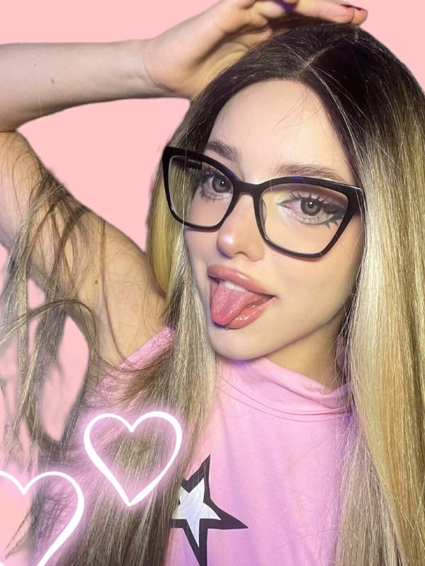Close-up selfie of a young woman with blonde hair and black glasses, posing with her tongue out and hand on her head against a pink background.