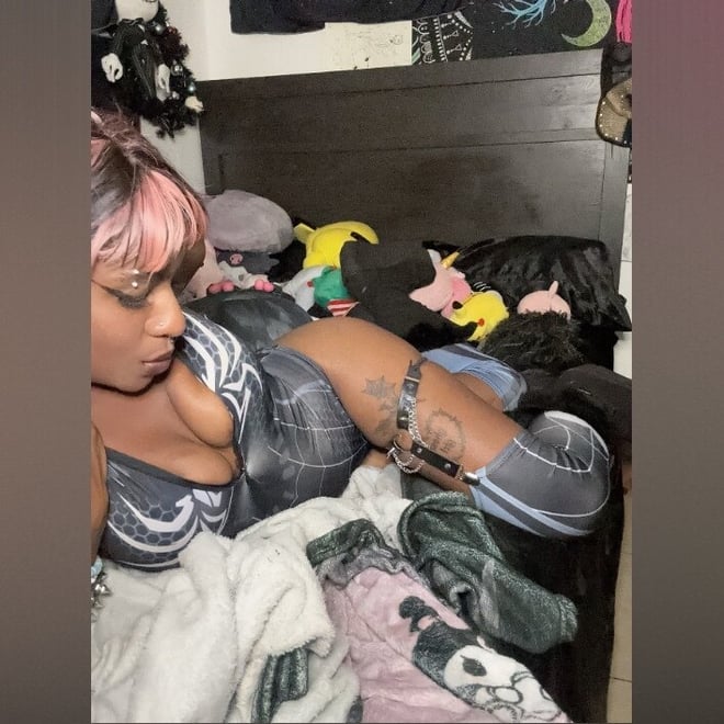 A woman with pink hair lounging on a bed in a gothic-themed bedroom surrounded by dark decor and stuffed animals.