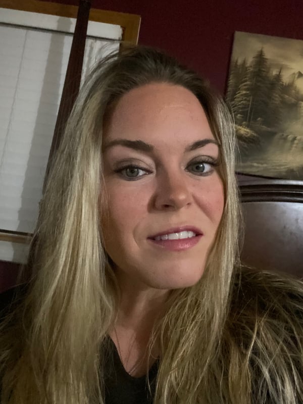 A smiling woman with long blonde hair poses indoors against a rustic wooden background with framed artwork.