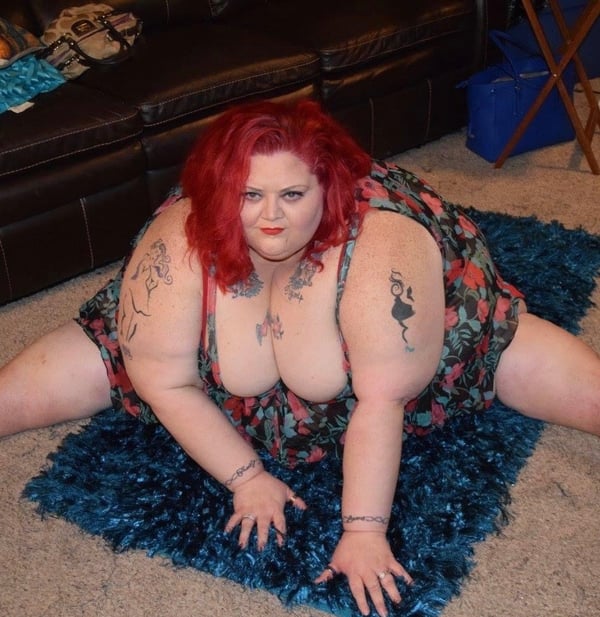 A woman with red hair sits on a blue rug in a living room, wearing a colorful floral dress, surrounded by furniture and decor.