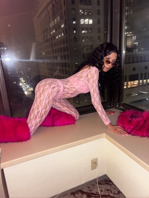 A woman posing on a windowsill in a high-rise room at night, wearing a lacy outfit and fluffy pink slippers, with city lights visible outside.