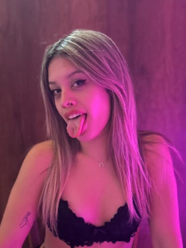 A young woman with long blonde hair smiling playfully with her tongue out, wearing lace top and necklace against a wooden wall with pink lighting.