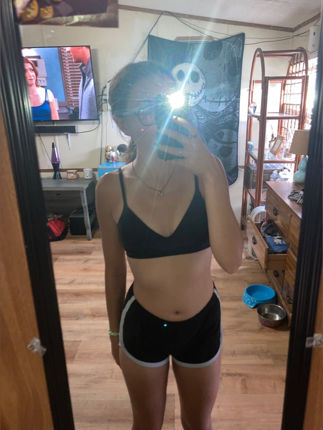 A young woman taking a mirror selfie in a black bralette and shorts inside a casual home with tapestries and furniture.